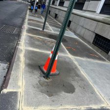 Cracked sidewalk slab around a sign pole base with a leaning cone